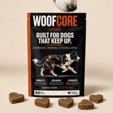 A stand-up pouch of WoofCore dog joint supplement placed on a beige surface, with several brown heart-shaped dog treats arranged around it and part of a dog's snout visible at the top edge. The package is black with orange side panels and white/orange branding, and the front shows a dog illustration with glowing orange joint highlights.